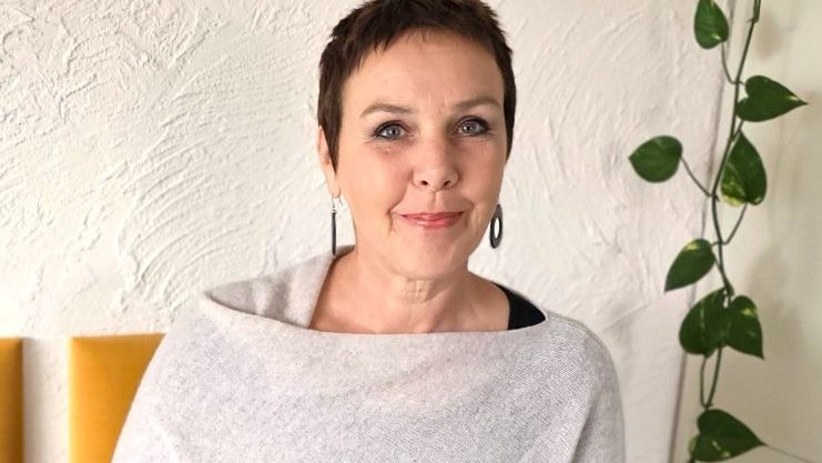 Über mich Frau mit kurzer Frisur, lächelt, trägt graue Kleidung und Ohrringe, Pflanzen im Hintergrund.