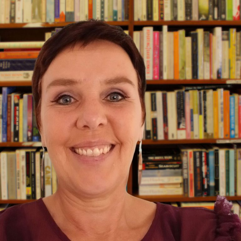 Frau mit kurzer Frisur, lächelt, trägt graue Kleidung und Ohrringe, Pflanzen im Hintergrund.