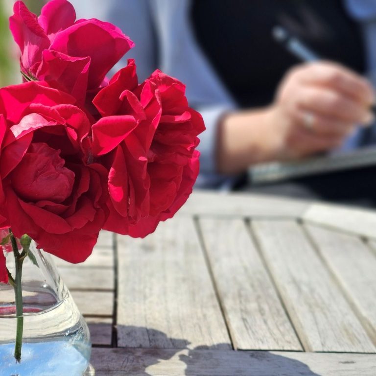 Karen schreibend im Grünen Frau sitzt an einem Tisch mit Blumenvase in einer grünen Feldlandschaft.