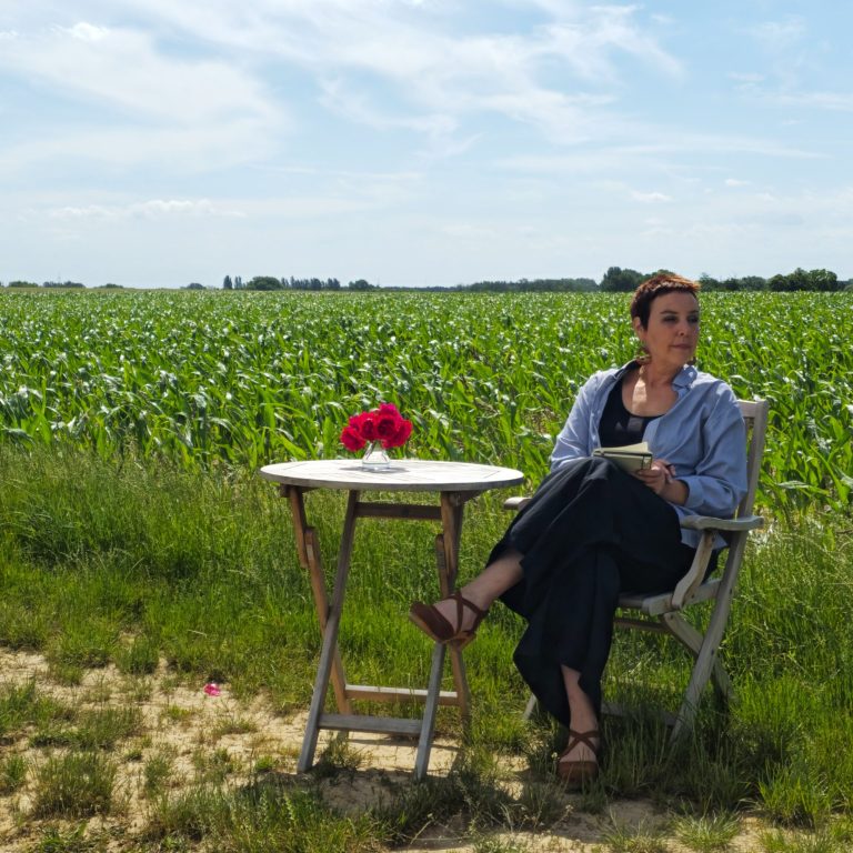 Karen schreibend im Grünen Frau sitzt an einem Tisch mit Blumenvase in einer grünen Feldlandschaft.